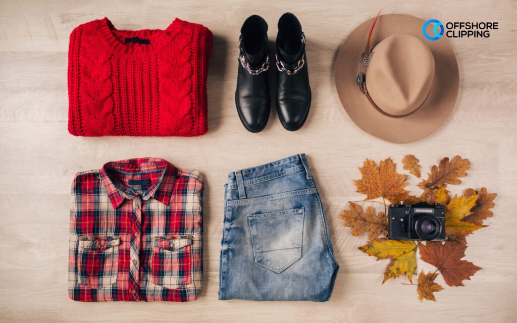 A flat lay collage featuring a red sweater, plaid shirt, blue jeans, black ankle boots, a beige hat with a feather, and a vintage camera, styled alongside autumn leaves on a wooden surface