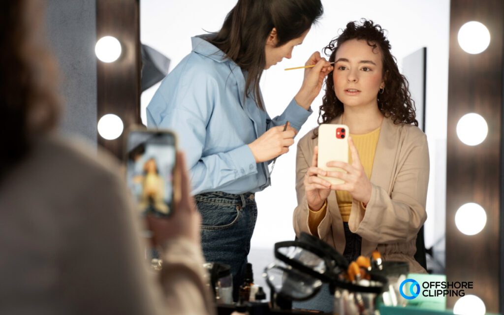 Makeup artist applying cosmetics to a model during a feature shot, with the model holding a smartphone in a lit studio setup.