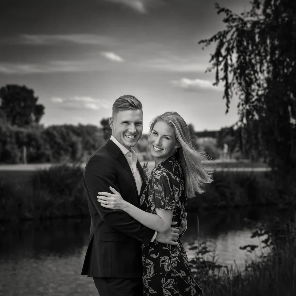 A smiling couple embraces beside a lake in a dramatic black-and-white outdoor portrait.