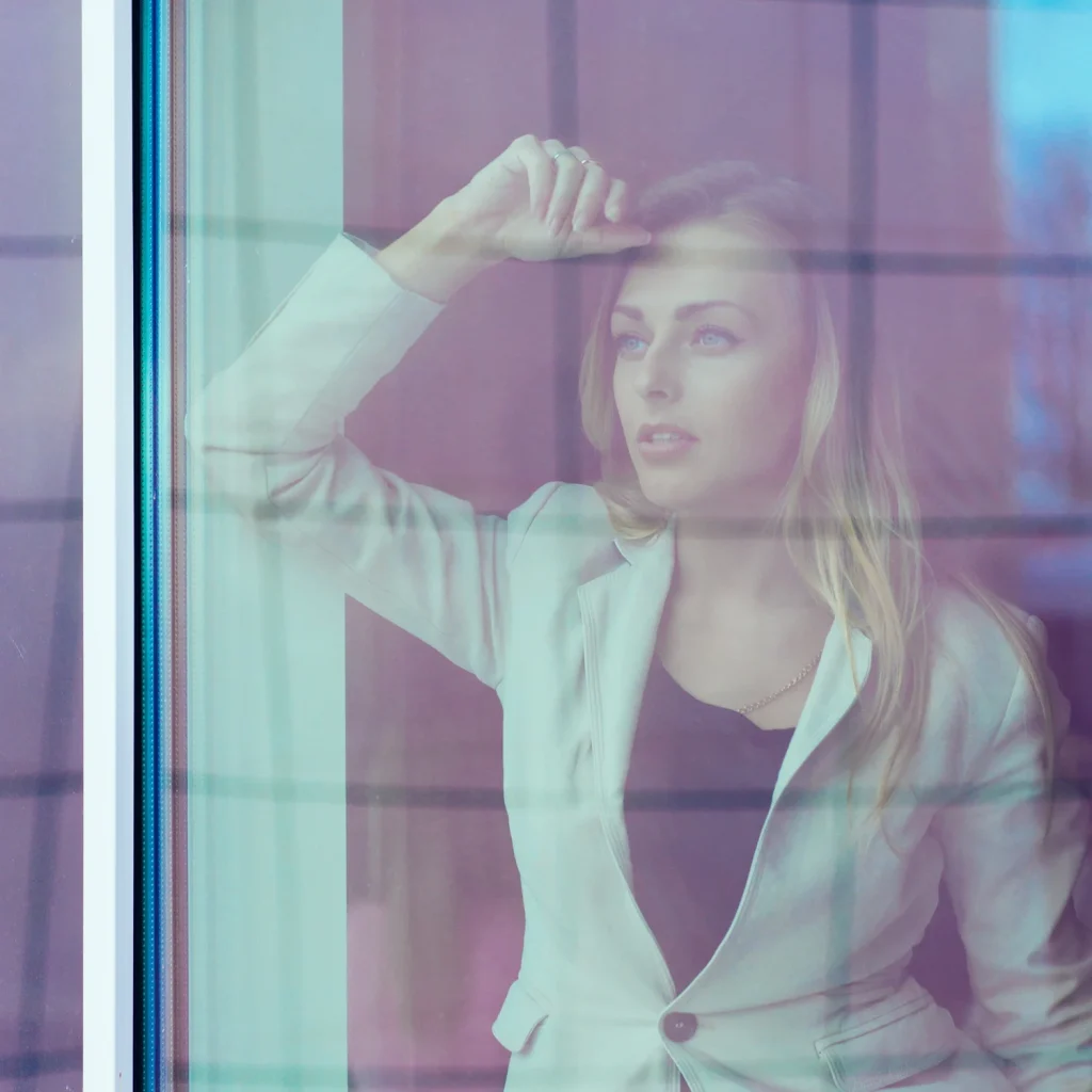 A softly blurred blonde woman in a light blazer looks outside through a reflective glass window.