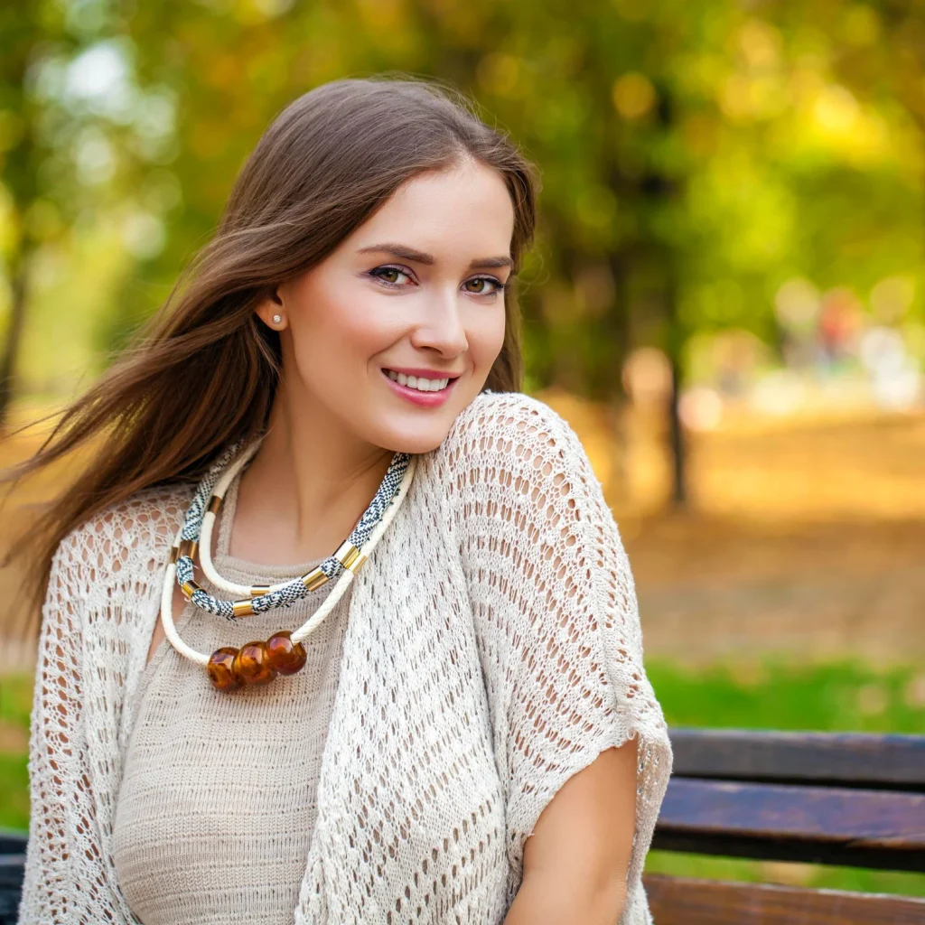 A smiling brunette woman in a cream knitted outfit poses outdoors on a park bench.