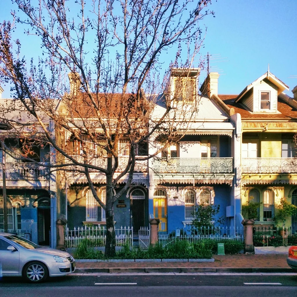 A row of colorful Victorian-style houses stands beneath a clear blue sky with parked cars outside.