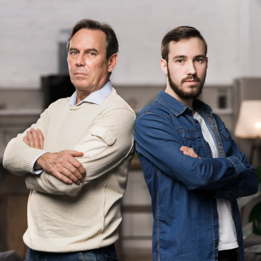 An older man and a younger bearded man stand back-to-back with folded arms in a modern interior.