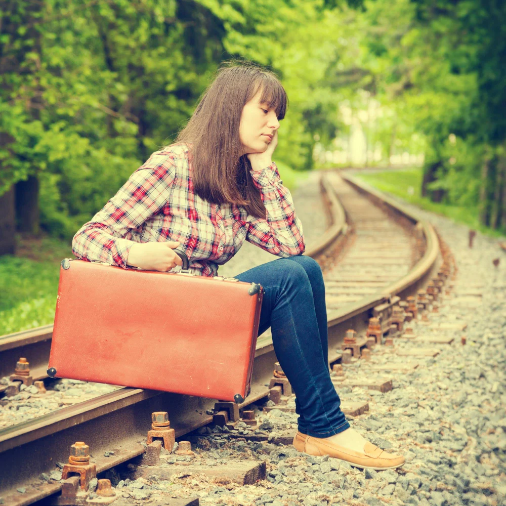 A young woman sits beside railway tracks with a large red suitcase in a green wooded area.