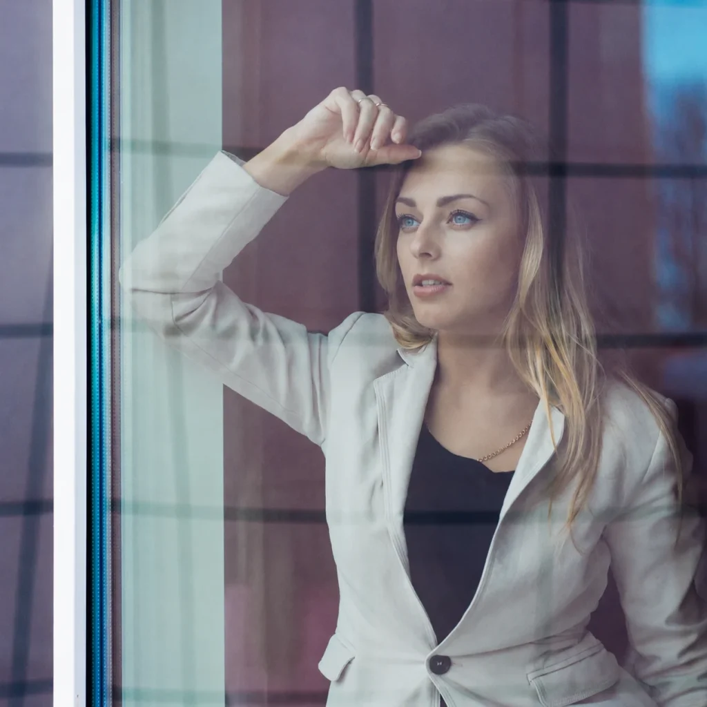 A blonde woman in a light blazer stands behind a window and looks into the distance.