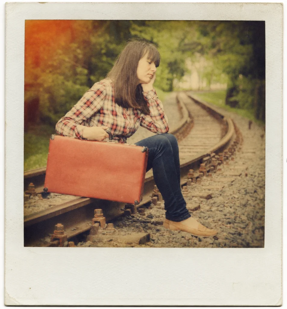 A vintage Polaroid-style photo shows a young woman sitting beside railway tracks with a red suitcase.