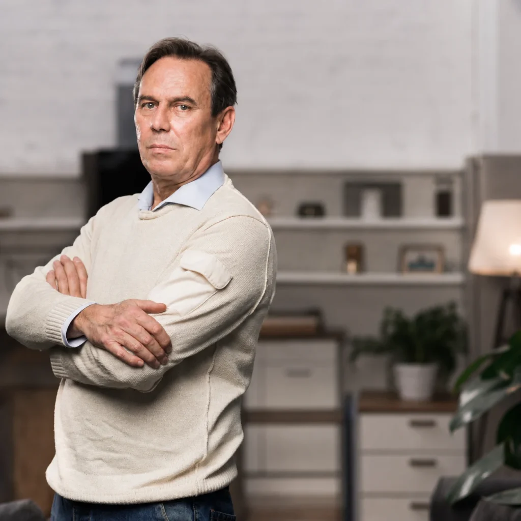 An older man wearing a cream sweater stands alone with folded arms in a softly lit interior.