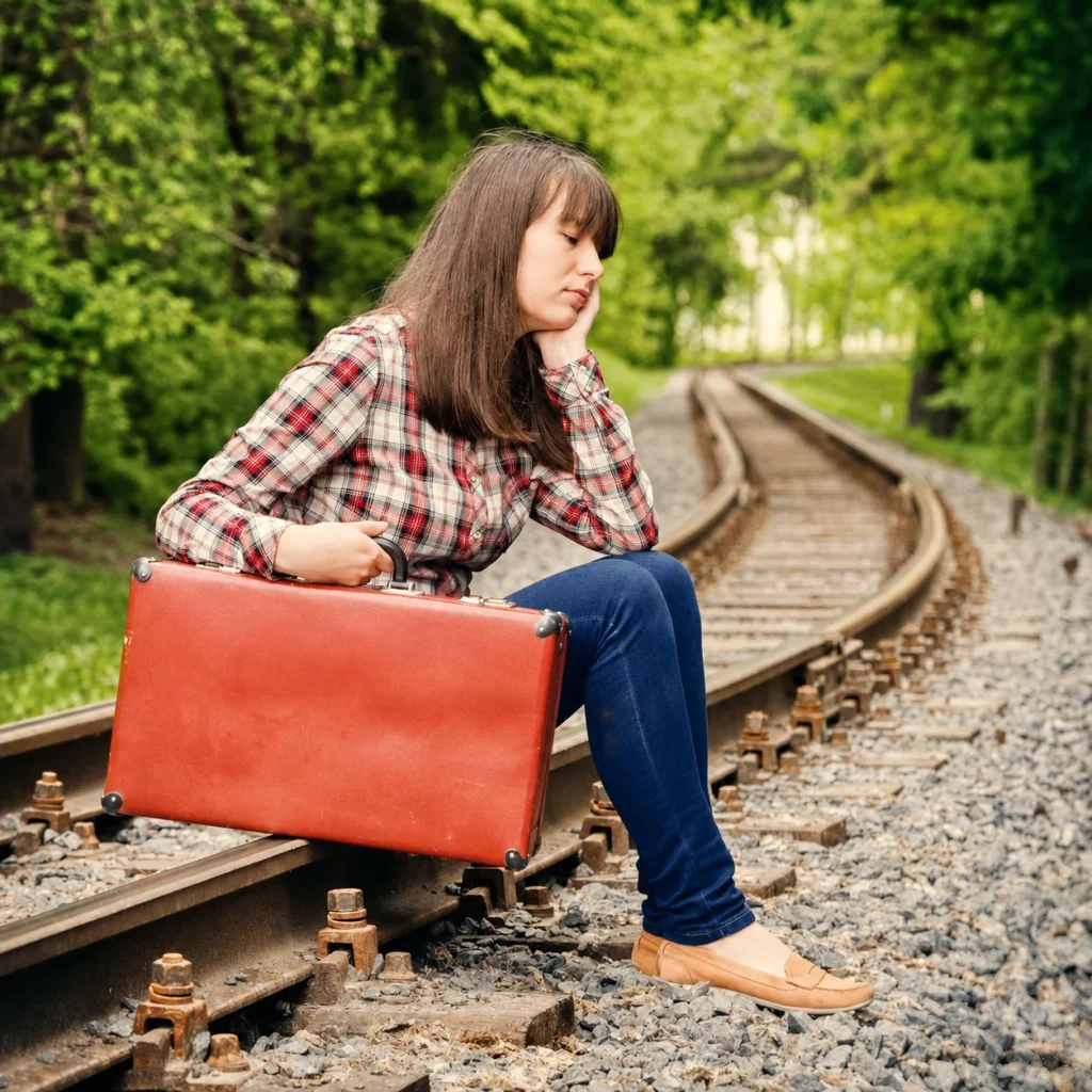 A young woman with a red suitcase sits beside railway tracks surrounded by green trees.