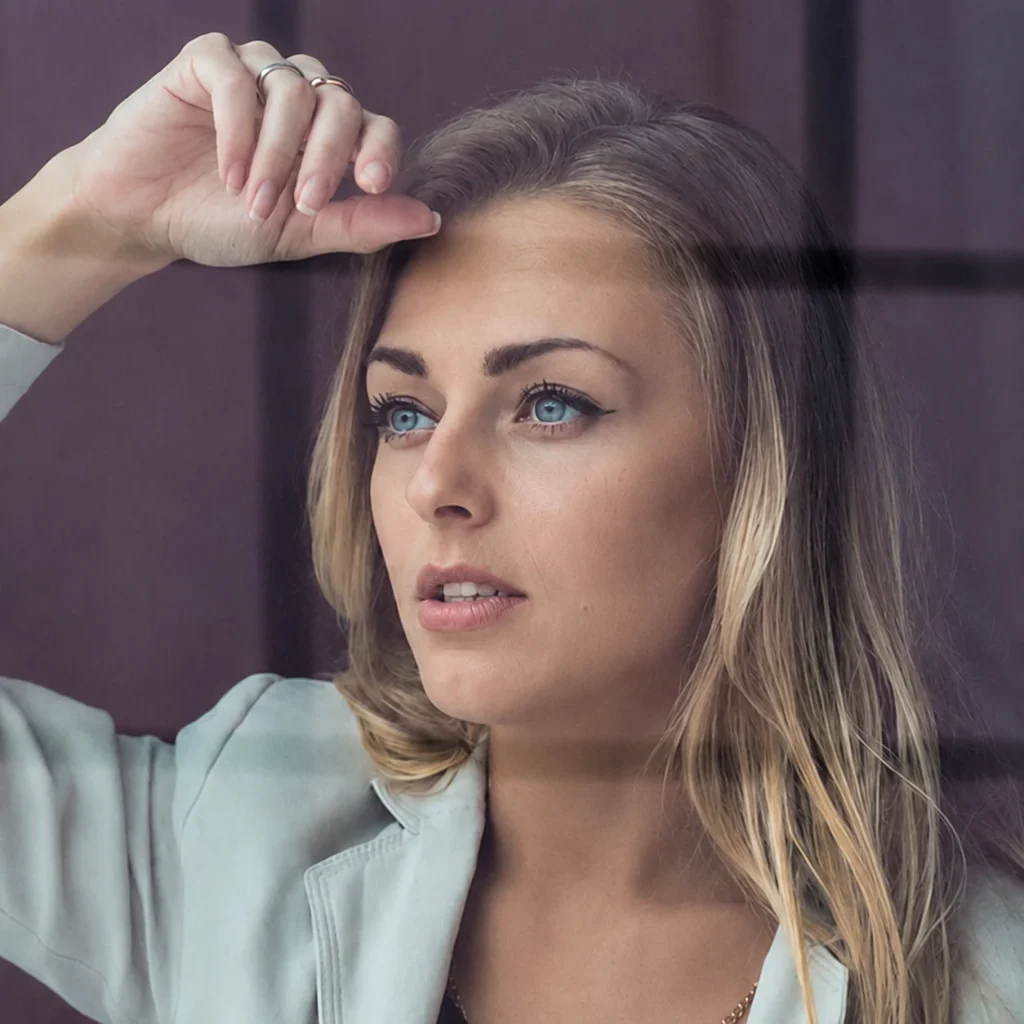 A blonde woman in a light blazer looks thoughtfully through a window in a sharp close-up portrait.
