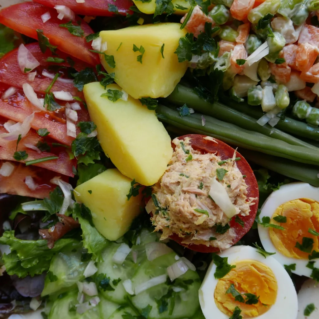 A close-up salad features potatoes, tomatoes, green beans, mixed vegetables, tuna, and boiled eggs.