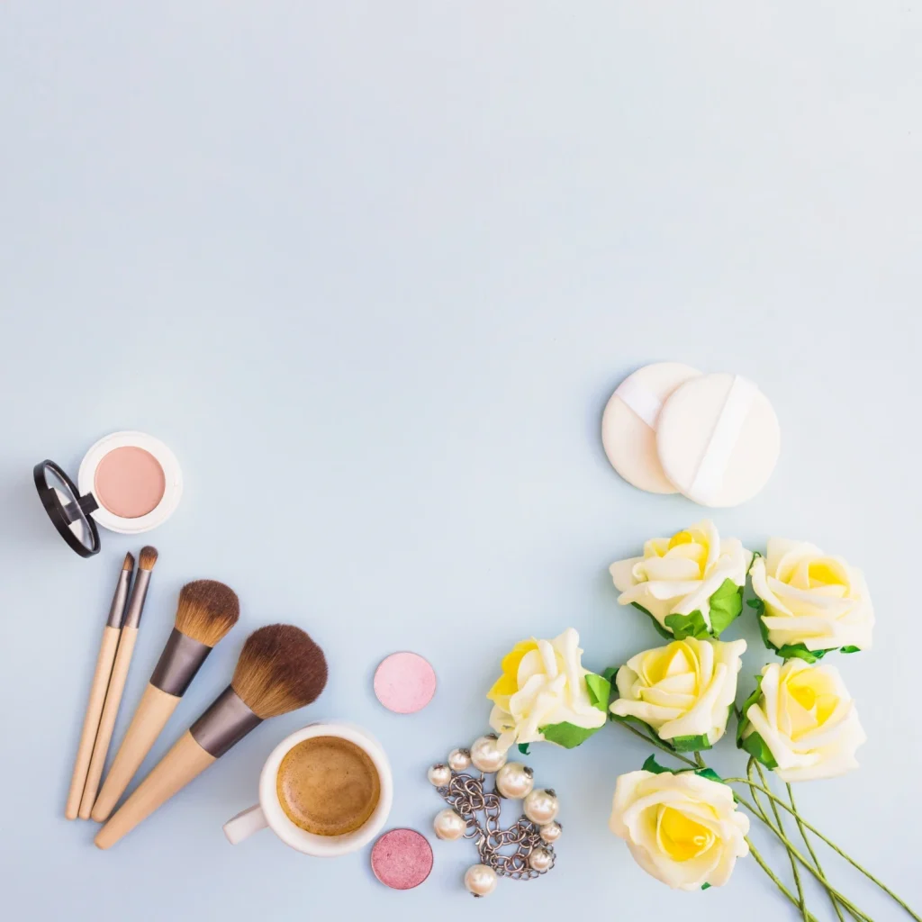Modern flat lay of beauty products, makeup brushes, coffee, jewelry, and flowers.