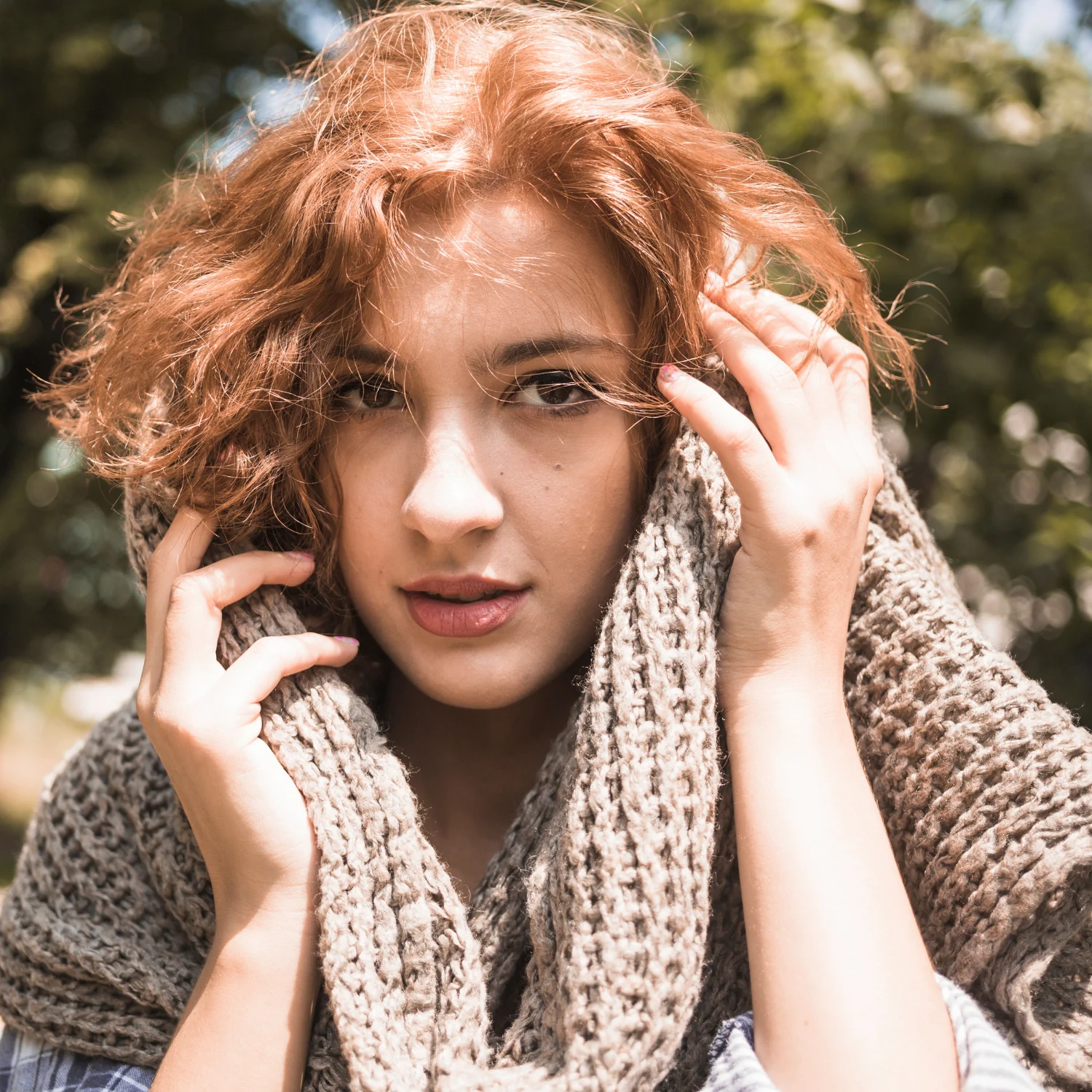 A red-haired woman wrapped in a chunky knitted scarf poses outdoors against a softly blurred background.