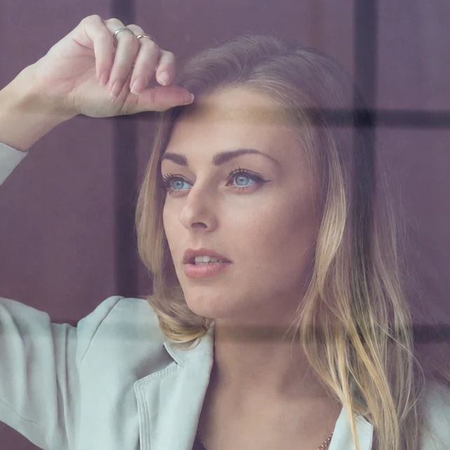 A blonde woman in a light blazer looks thoughtfully through a reflective glass window.
