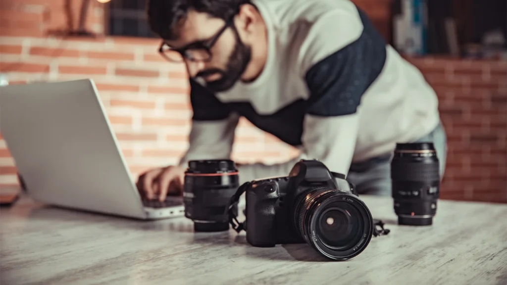 Photographer working on a laptop beside camera and lenses, representing specialized outsourced editing skills
