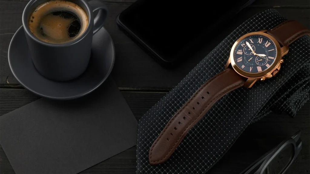 Rose gold luxury chronograph watch with brown leather strap styled with coffee and accessories on dark background