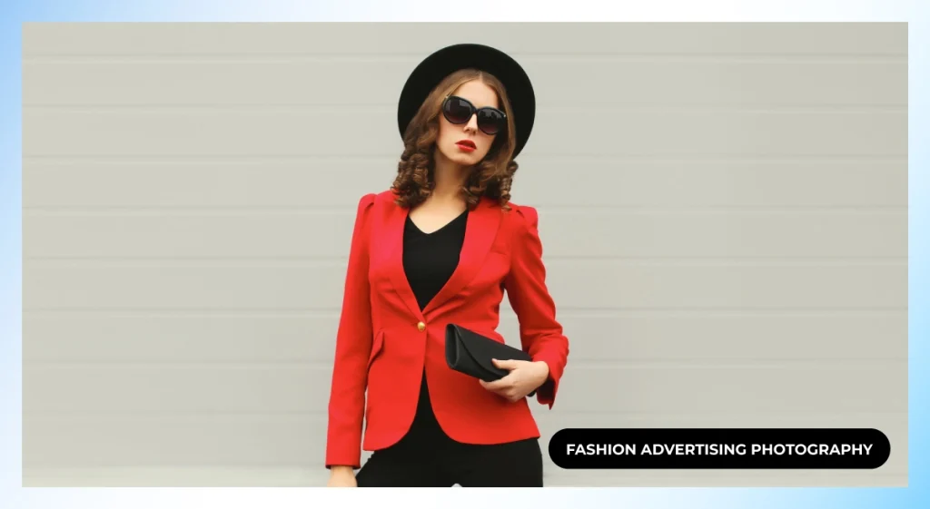 Model in a red blazer, black hat and sunglasses holding a black clutch against a gray wall, fashion advertising photography